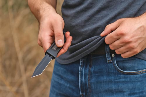 Man deploying a black automatic knife outdoors while holding his shirt.