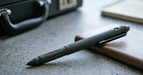 A discreet tactical pen resting next to a notebook on a gray textured surface