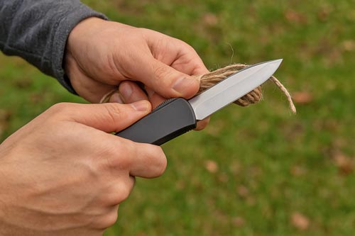 Hands cutting a bundle of twine with a black automatic knife outdoors.