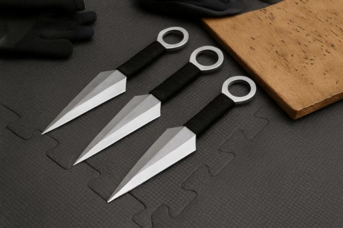 Three stainless steel kunai knives on a black training mat near a wooden target.