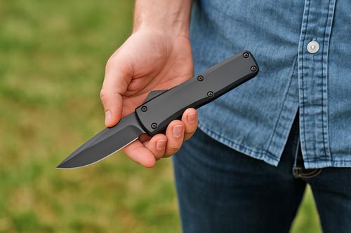 Person holding a black automatic knife outdoors while wearing a blue denim shirt.