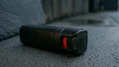 A black pepper spray canister with a red safety flip-top on stone.