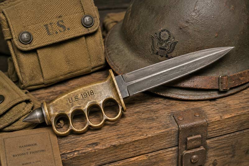Historical U.S. Model 1918 Mark I trench knife with brass knuckle guard displayed beside a World War I helmet and military gear.